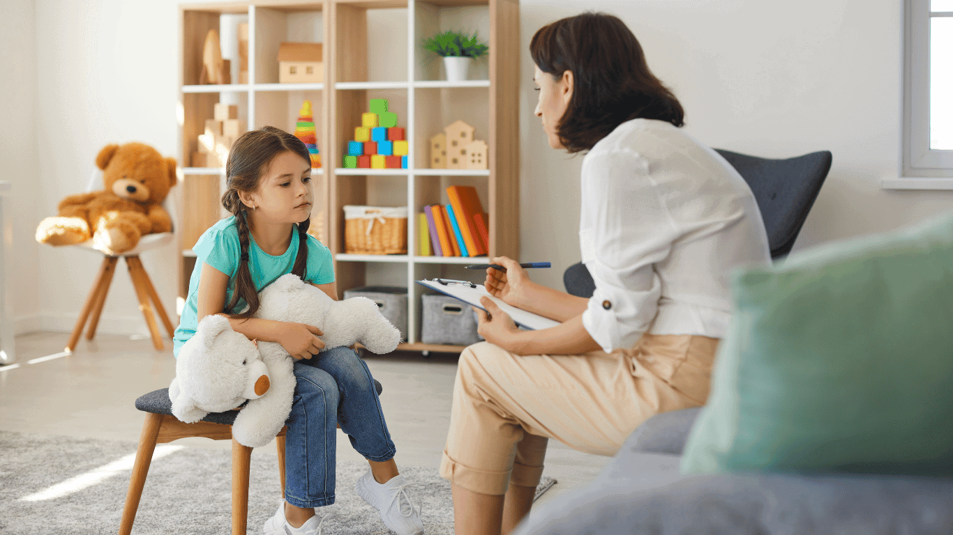 A young girl holding a stuffed animal sits across from an adult woman with a clipboard, having a serious conversation in a bright room with toys and shelves in the background.