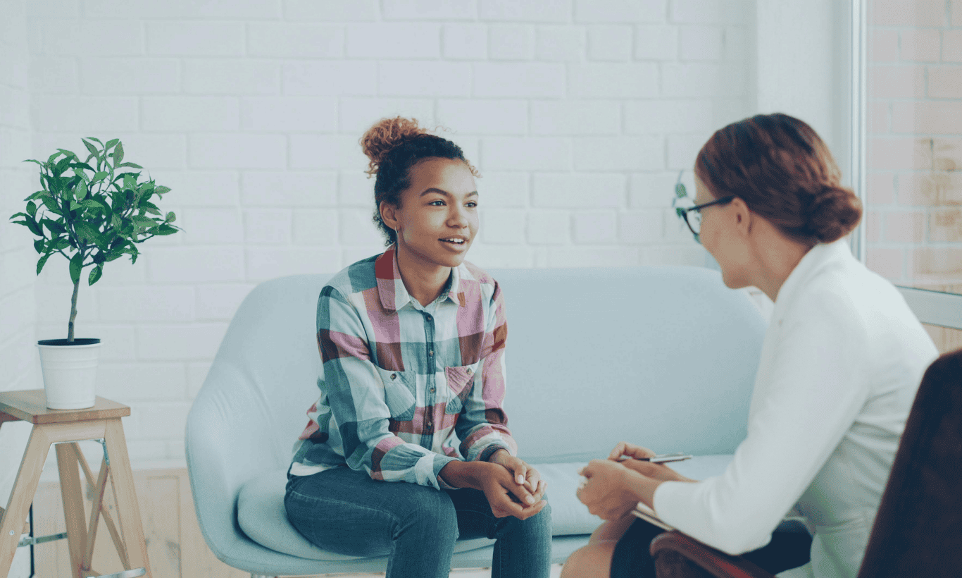 Two women sit on a light blue couch in conversation. One wears a plaid shirt and faces the other, who holds a notepad and pen. A potted plant is on a small table nearby.