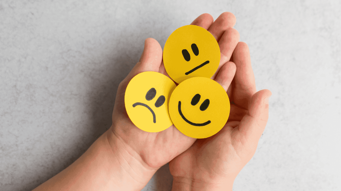 A pair of hands holds three yellow paper circles with drawn faces: one happy, one sad, and one neutral, representing different emotions.