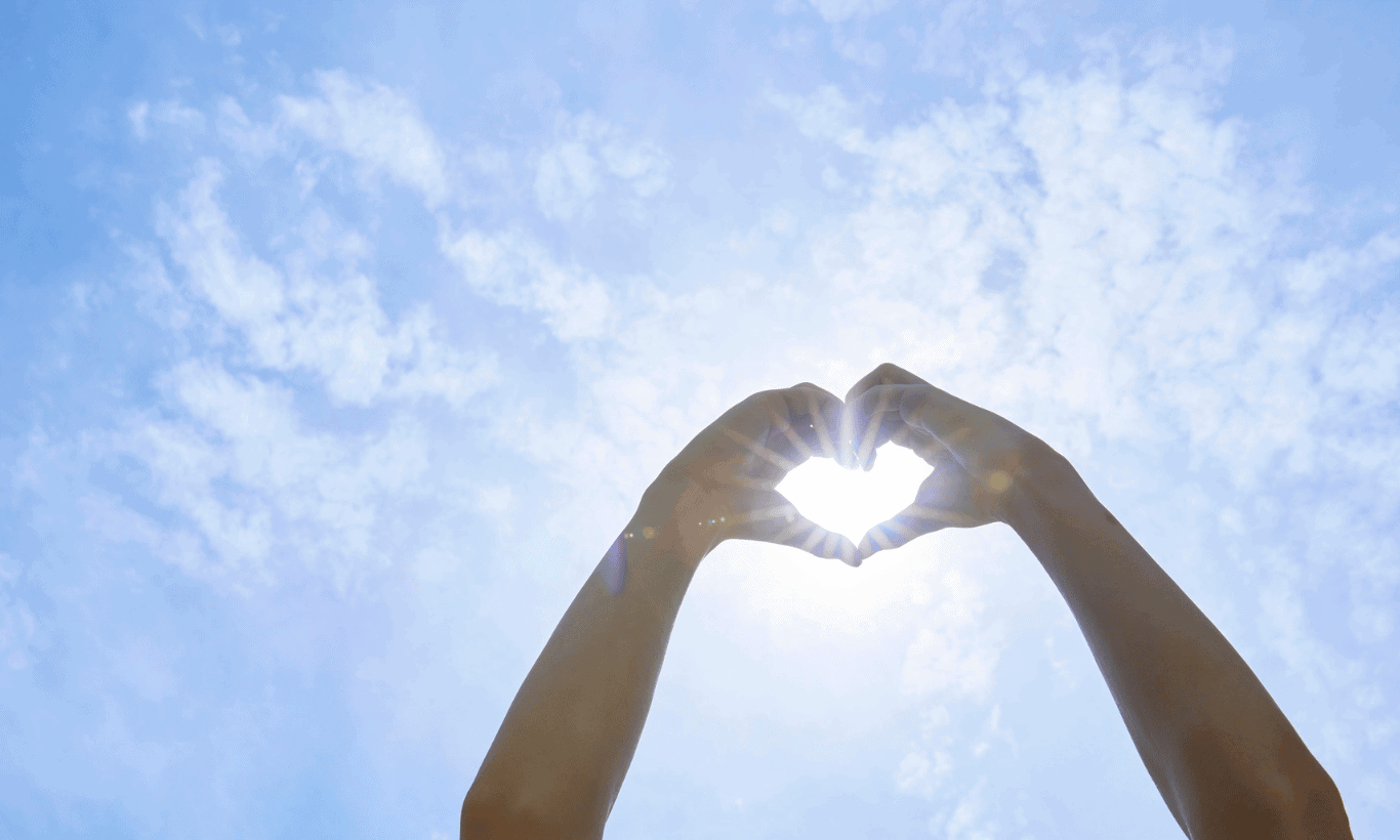 Two hands held up against a bright blue sky form the shape of a heart, with sunlight shining through the heart shape and scattered clouds in the background.