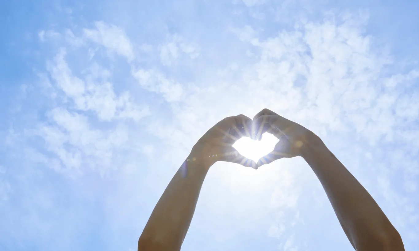 Two hands held up against a bright blue sky form the shape of a heart, with sunlight shining through the heart shape and scattered clouds in the background.