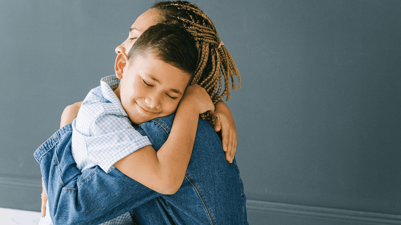 A young boy with short hair smiles with eyes closed while hugging an adult wearing a denim jacket, standing in front of a plain gray wall.