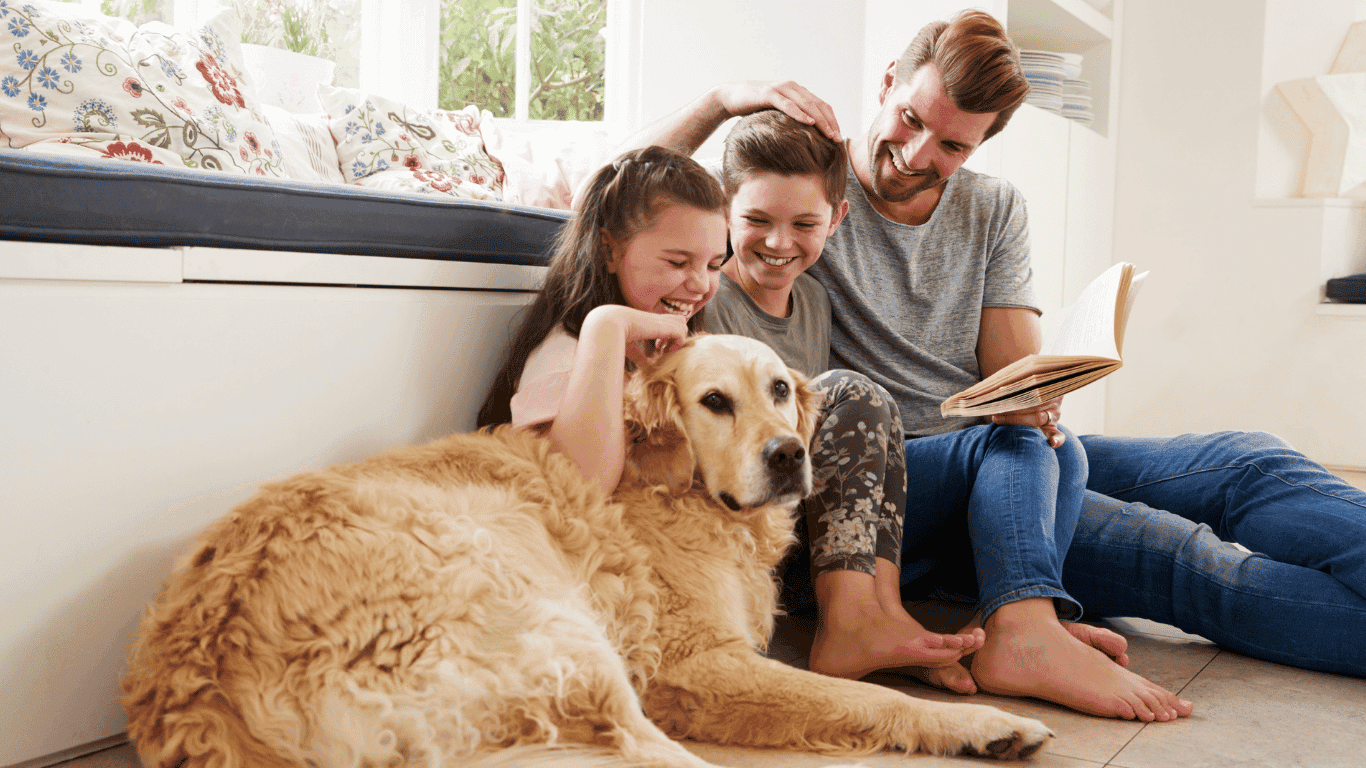 A man and two children sit on the floor, smiling and petting a golden retriever. The man is holding an open book, and they appear happy and relaxed in a cozy, well-lit room.
