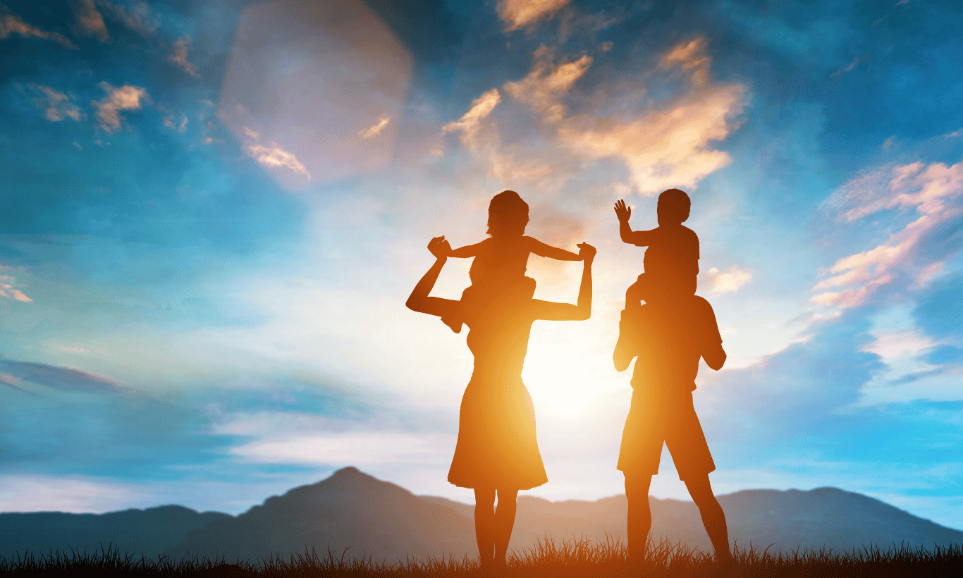 Silhouette of two adults carrying two children on their shoulders, standing outdoors at sunset with mountains and a vibrant sky in the background.
