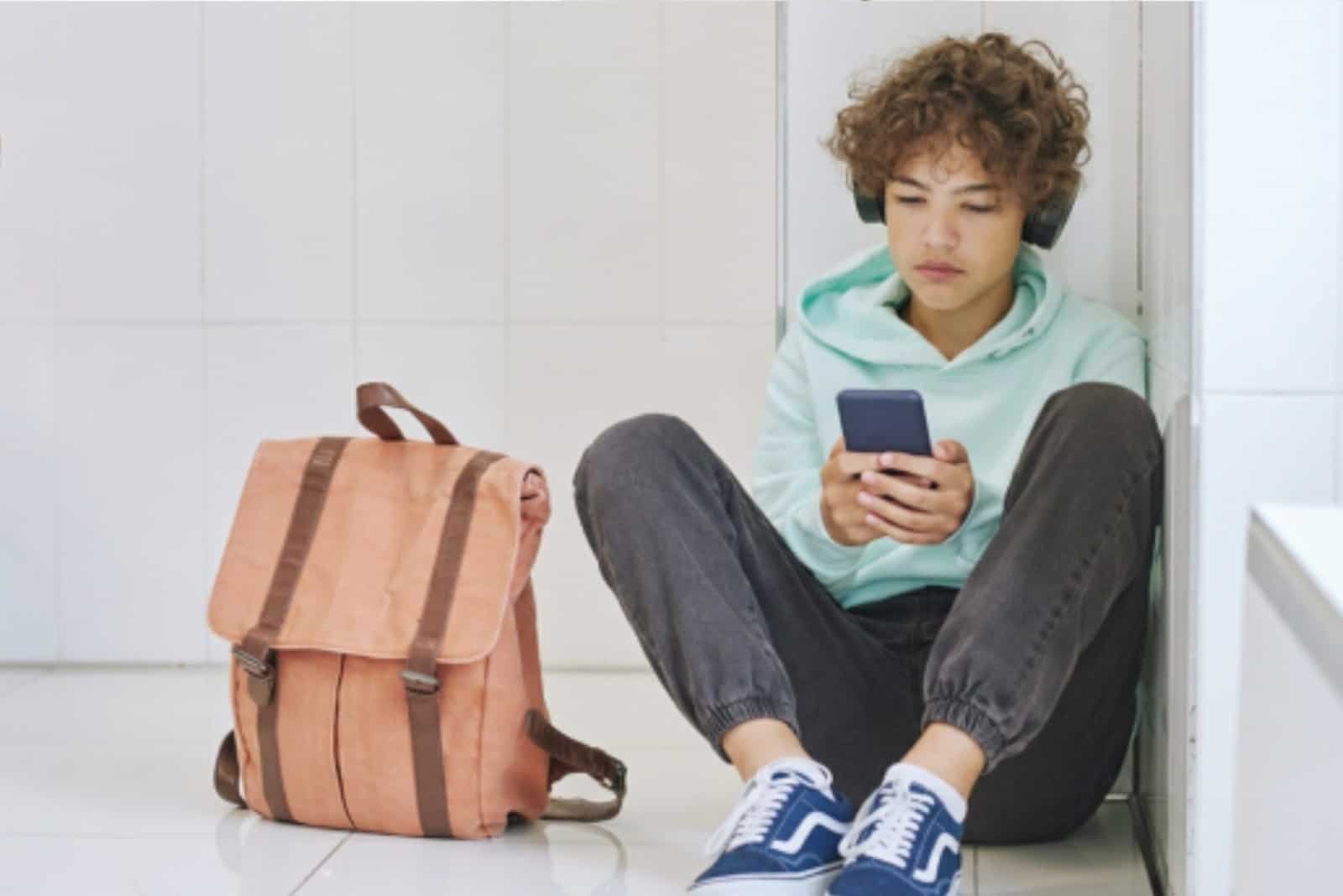 Teen with curly hair, wearing headphones, a mint hoodie, and blue sneakers, sits on the floor using a smartphone. A pink backpack is beside them against a white tiled wall.
