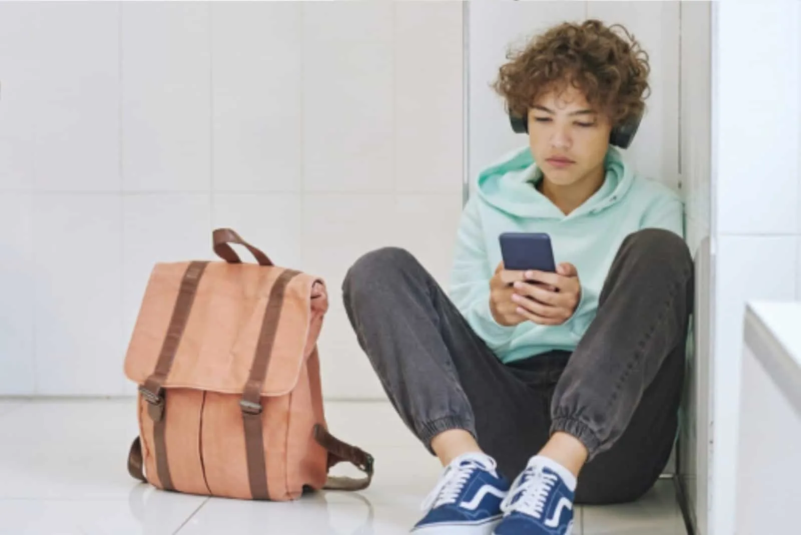 Teen with curly hair, wearing headphones, a mint hoodie, and blue sneakers, sits on the floor using a smartphone. A pink backpack is beside them against a white tiled wall.