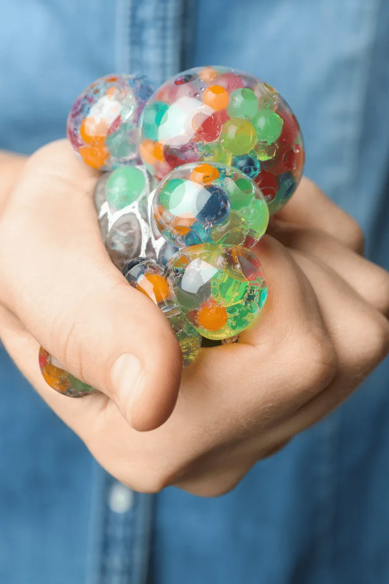 A hand squeezes a clear stress ball filled with colorful beads and gel, set against a blurred background of denim fabric.