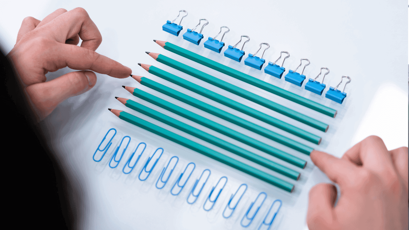 Two hands point at six green pencils neatly lined up on a white surface, surrounded by blue paper clips on the left and binder clips on top, all arranged in parallel rows.