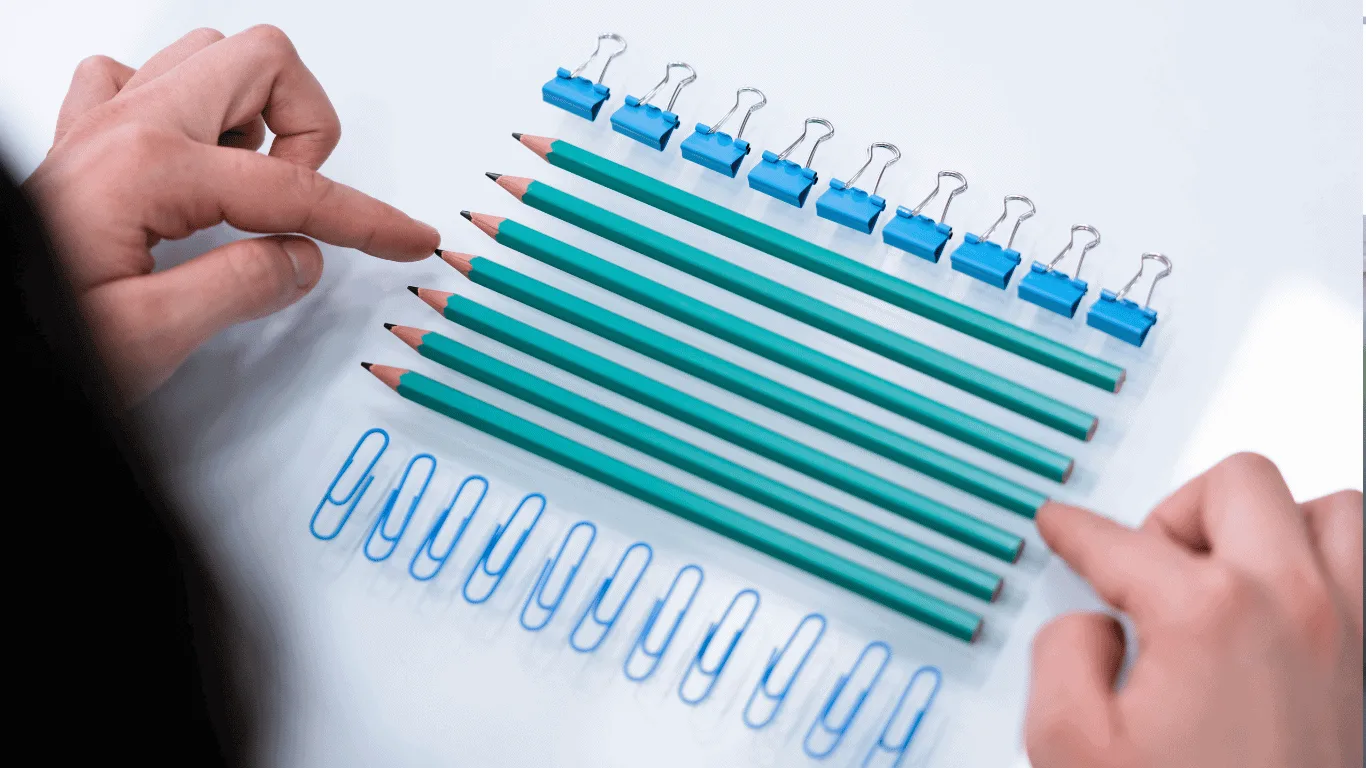 Two hands point at six green pencils neatly lined up on a white surface, surrounded by blue paper clips on the left and binder clips on top, all arranged in parallel rows.