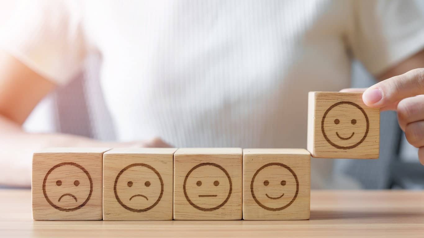 A hand lifts a wooden block with a happy face from a row of blocks showing faces from sad to happy, representing different emotions.