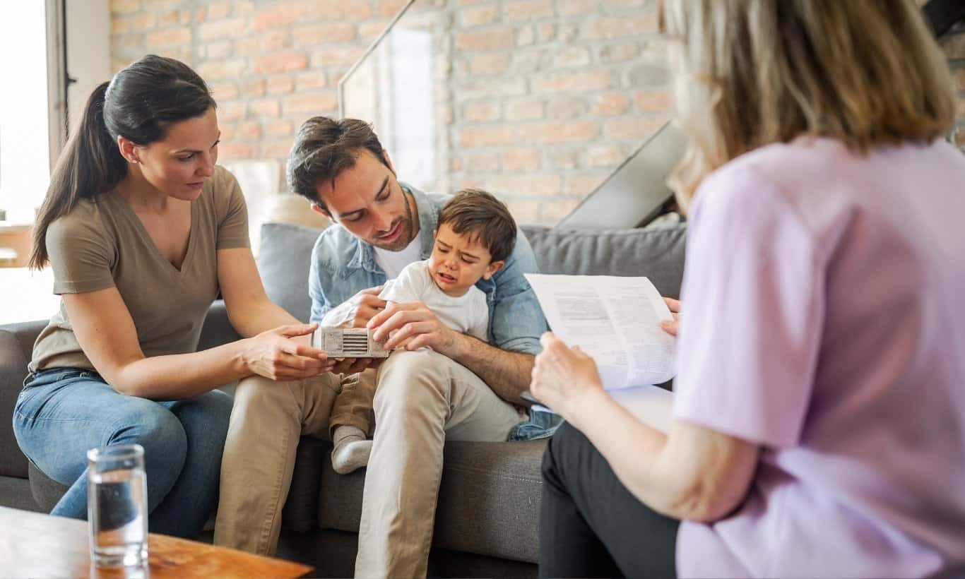 A couple comforts their upset young child on a couch during a therapy or counseling session, while a professional with papers sits across from them.