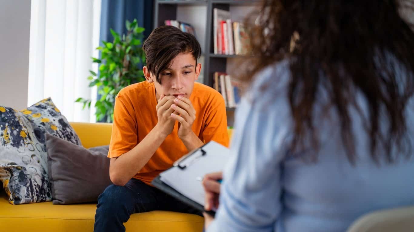 A teenage boy in an orange shirt sits on a yellow couch, looking thoughtful and anxious, while a person with a clipboard, likely a therapist, sits across from him in an office with bookshelves.