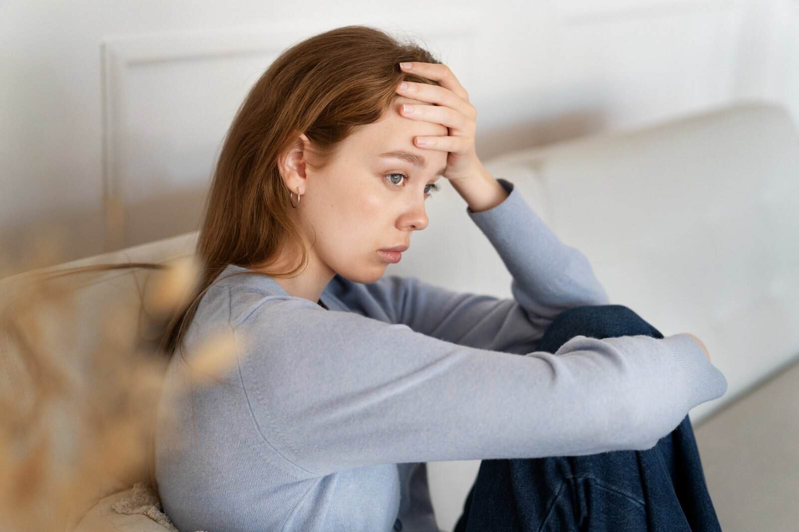 A young woman sits on a couch, looking stressed and worried, with her hand resting on her forehead and her knees drawn up toward her chest.