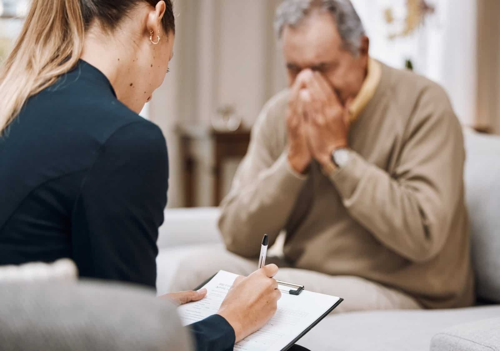 A woman with a clipboard listens to an older man sitting on a couch, who covers his face with his hands, appearing upset.