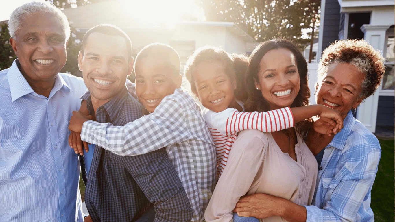 Three generations of a smiling family stand closely together outside on a sunny day, with two children embracing the adults. Everyone appears happy and joyful, and the background shows houses and trees.