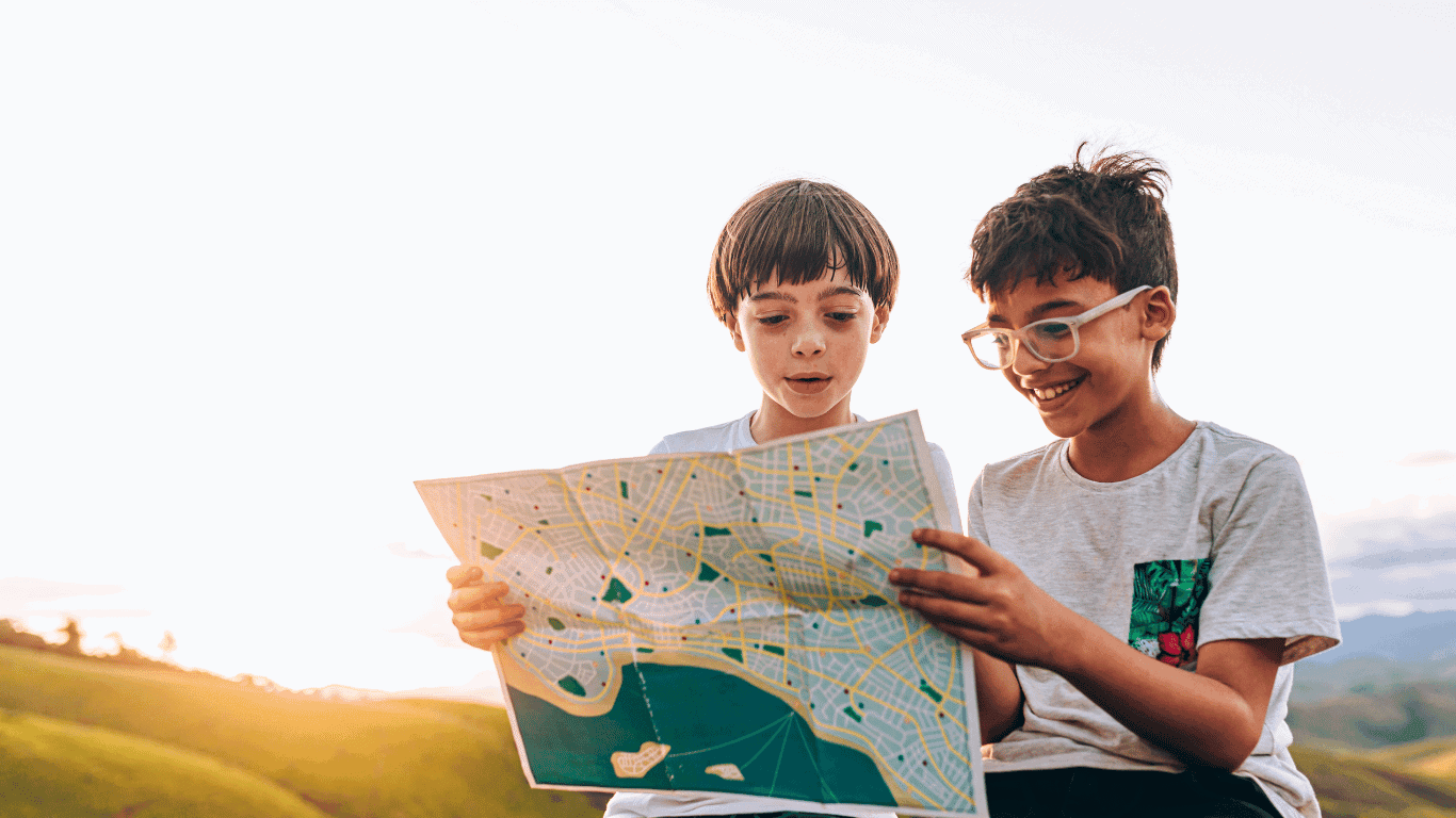 Two children sit outdoors on grassy hills at sunset, smiling and looking at a large paper map together. One child wears glasses and a t-shirt; the other has a bowl haircut and a white shirt.