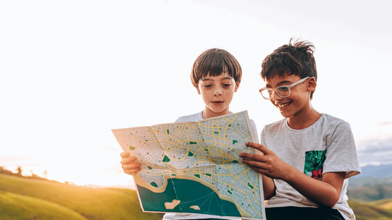 Two children sit outdoors on grassy hills at sunset, smiling and looking at a large paper map together. One child wears glasses and a t-shirt; the other has a bowl haircut and a white shirt.