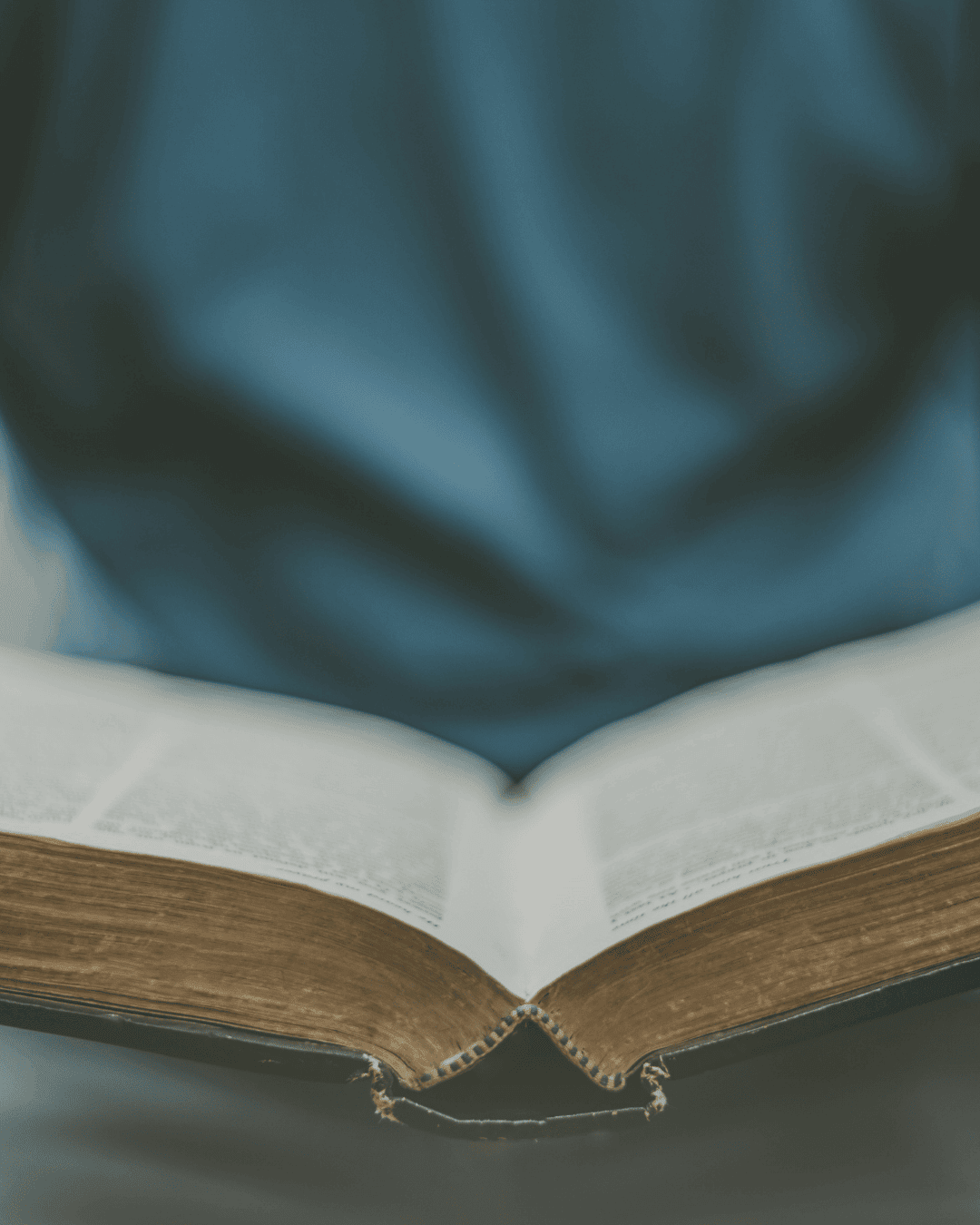 A person wearing a blue shirt is sitting with an open book, possibly a Bible, on their lap. The focus is on the books pages, while the persons upper body is blurred in the background.
