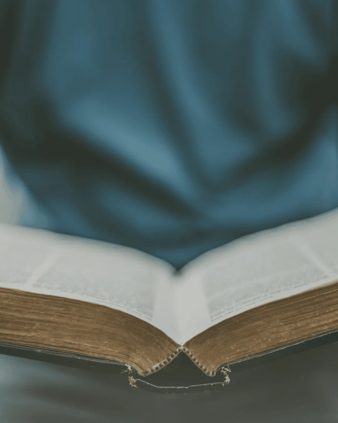 A person wearing a blue shirt is sitting with an open book, possibly a Bible, on their lap. The focus is on the books pages, while the persons upper body is blurred in the background.