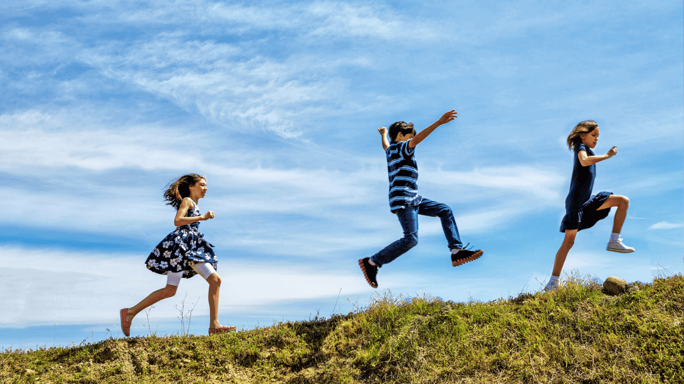 Three children are running and jumping up a grassy hill under a blue sky with scattered clouds.