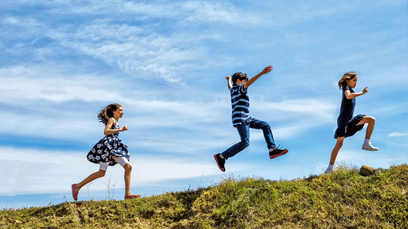 Three children are running and jumping up a grassy hill under a blue sky with scattered clouds.