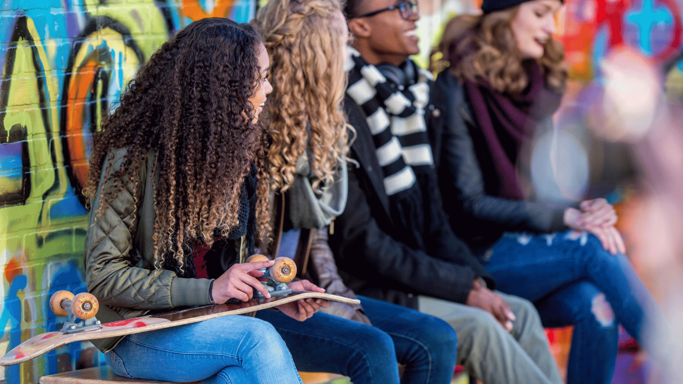 Four young people sit in front of a colorful graffiti wall; one holds a skateboard. They are dressed in casual, warm clothes and appear to be talking and enjoying each others company.