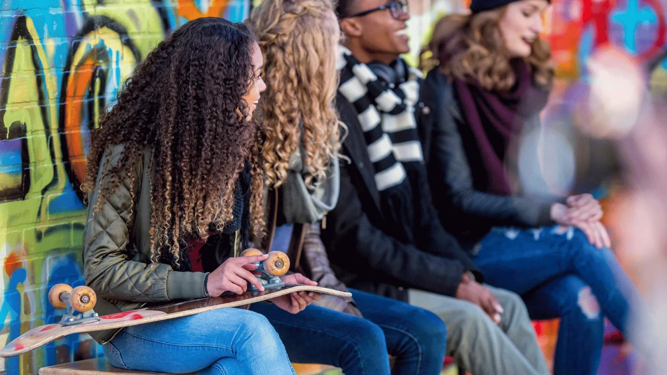 Four young people sit in front of a colorful graffiti wall; one holds a skateboard. They are dressed in casual, warm clothes and appear to be talking and enjoying each others company.