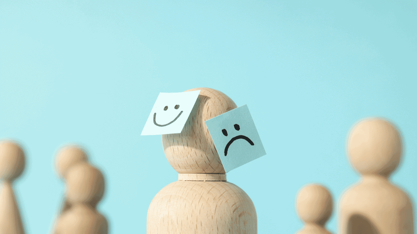 A wooden figure with a sad face sticky note on its front and a happy face sticky note on its side stands among other plain wooden figures against a blue background.