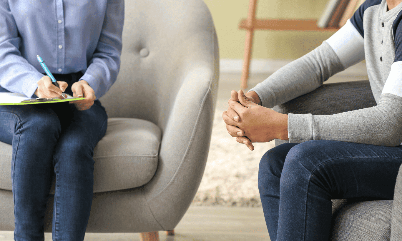 Two people sit in chairs facing each other, one taking notes on a clipboard and the other with hands clasped, suggesting a therapy or counseling session in a neutral setting.