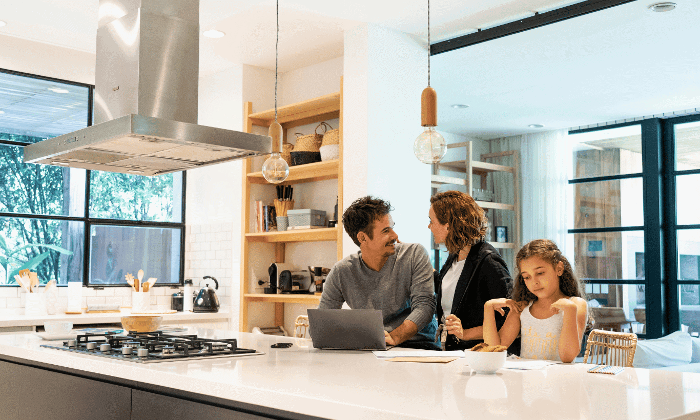 A man and woman talk at a kitchen island with a laptop and papers while a young girl eats cereal nearby. Bright, modern kitchen with large windows and shelves in the background.
