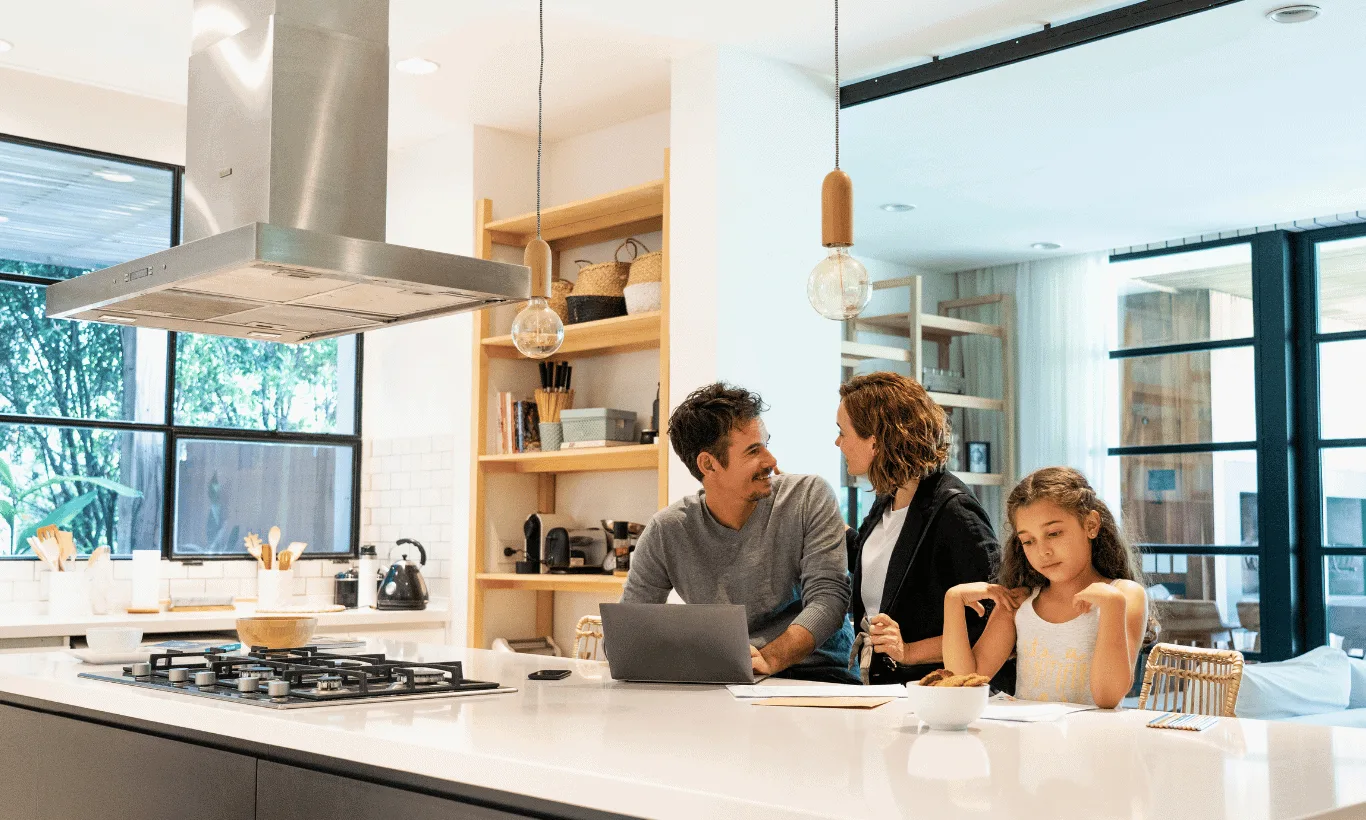 A man and woman talk at a kitchen island with a laptop and papers while a young girl eats cereal nearby. Bright, modern kitchen with large windows and shelves in the background.
